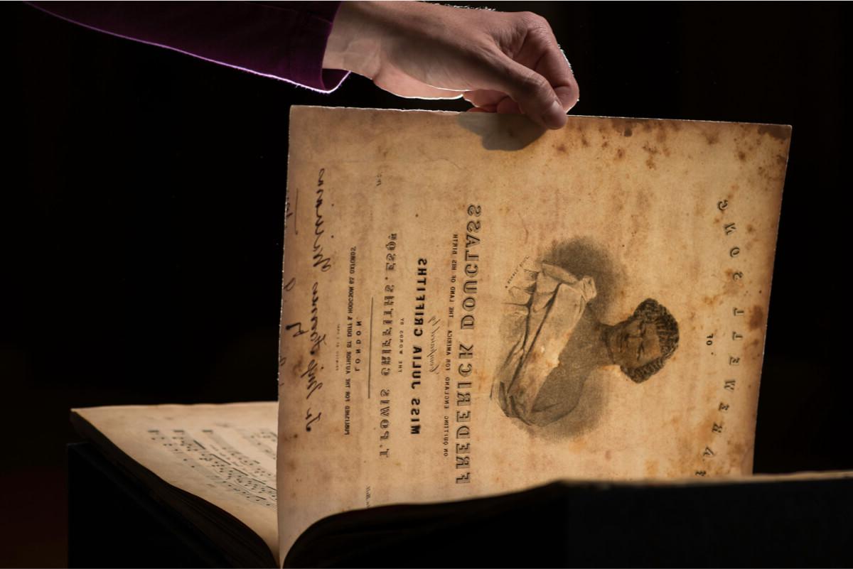 a hand holds up a sheet of music with an image of Frederick Douglass and the title FAREWELL SONG TO FREDERICK DOUGLASS 